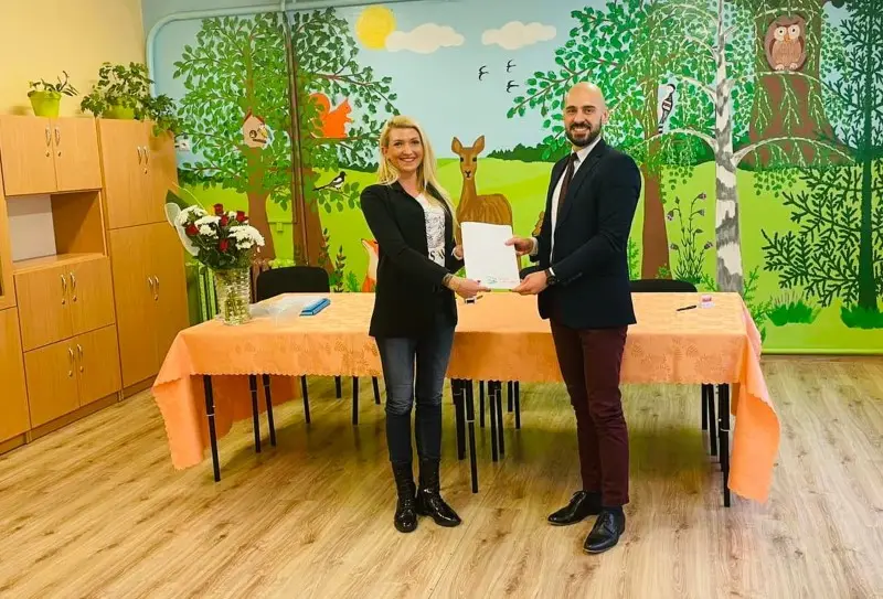 Zdjęcie o orientacji poziomej przedstawia kobietę i mężczyznę stojących wewnątrz kolorowej sali przedszkolnej, trzymających wspólnie biały dokument. Kobieta po lewej stronie ma na sobie czarną marynarkę, dżinsy i czarne botki, natomiast mężczyzna po prawej jest ubrany w granatową marynarkę, bordowe spodnie i eleganckie buty. Para znajduje się przed długim stołem nakrytym brzoskwiniowym obrusem, na którym stoi wazon z bukietem czerwonych róż. Tło stanowi ozdobna ściana pomalowana w motywy leśne z wizerunkami drzew, sarny, sowy oraz roślinności. Po lewej stronie kadru widoczna jest wysoka, drewniana szafka z drzwiczkami, a całe wnętrze utrzymane jest w ciepłej, przyjaznej dla dzieci stylistyce.