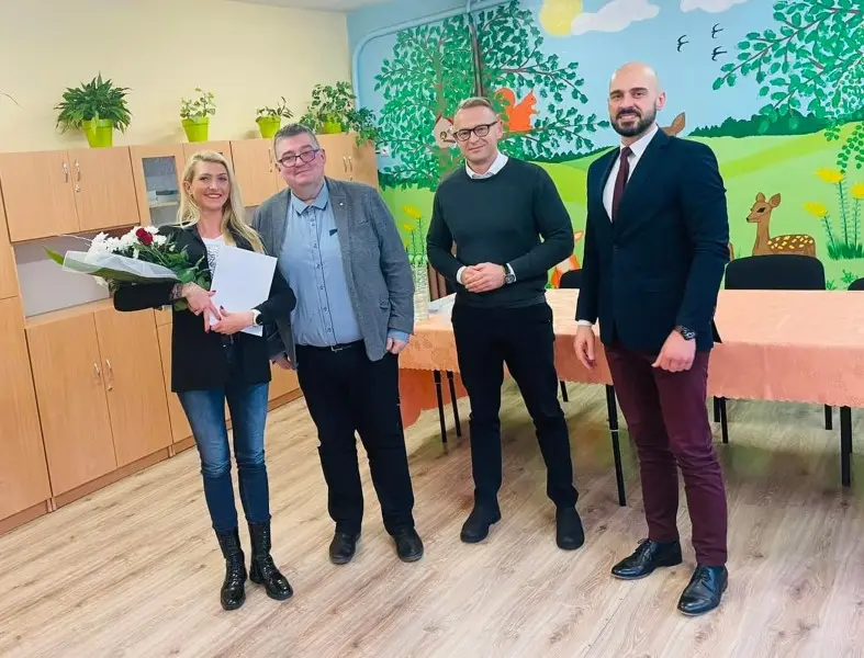 Zdjęcie w orientacji poziomej przedstawia grupę czterech osób pozujących w jasnym wnętrzu sali przedszkolnej lub edukacyjnej. Po lewej stronie stoi kobieta z blond włosami, trzymająca bukiet kwiatów oraz białą teczkę, ubrana w ciemną marynarkę, jeansy i wysokie czarne buty. Obok niej znajduje się trzech mężczyzn w eleganckich strojach: pierwszy w szarej marynarce i koszuli, drugi w ciemnym swetrze, a trzeci w granatowej marynarce z bordowym krawatem. Tło stanowi ściana ozdobiona kolorowym malowidłem ściennym z motywem przyrody, lasu, jelonka i wiewiórki, oraz jasne drewniane szafki wzdłuż ściany. Przed grupą znajduje się stół nakryty brzoskwiniowym obrusem, a na podłodze widoczne są panele o fakturze jasnego drewna. Całość kompozycji ma charakter oficjalnego spotkania lub uroczystości w placówce oświatowej.